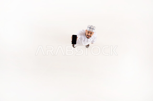 استخدام الأجهزة التقنية الحديثة في الحياة اليومية, النظر إلى الكاميرا بإيماءات السعادة والسرور, بورتريه علوي عامودي لرجل عربي خليجي عماني مبتسم يرتدي الدشداشة والكمة التقليدية يمسك بيده الهاتف المحمول, عرض شاشة سوداء فارغة عبر الجوال, خلفية بيضاء