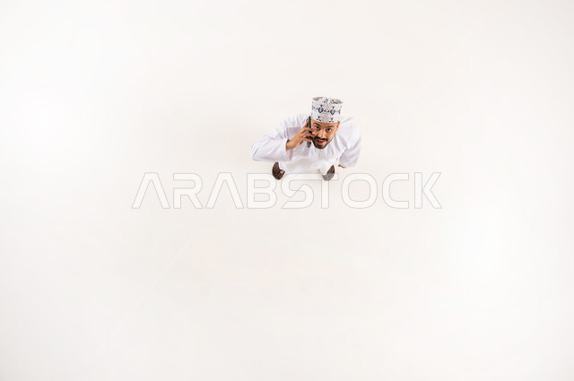 استخدام الأجهزة التقنية الحديثة في الحياة اليومية, النظر إلى الكاميرا بإيماءات السعادة والسرور, بورتريه علوي عامودي لرجل عربي خليجي عماني مبتسم يرتدي الدشداشة والكمة التقليدية يجري مكالمة هاتفية عبر الهاتف المحمول, استخدام الموبايل للتواصل والرد على الاتصال, خلفية بيضاء