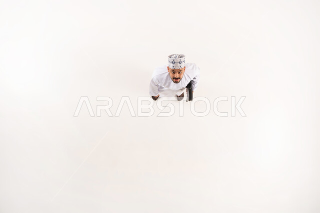 Concern for outward appearance, concept of masculinity and elegance, standing upright and upright, vertical overhead portrait of a smiling Omani Gulf Arab man wearing the traditional dishdasha and kummah, looking at the camera with expressions and gestures of pleasure and happiness, white background