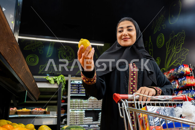 الاستمتاع بالتسوق في المولات والمراكز التجارية، امراة عربيه خليجيه اماراتية تقوم بشراء ثمار المانجو الطازجه، الاستفادة من العروض والخصومات، سيده سعودية محجبة ترتدي العباءة تحمل بيدها السلة، شراء المنتجات من السوبر ماركت، مخاصيل زراعيه وطنه سعوديه