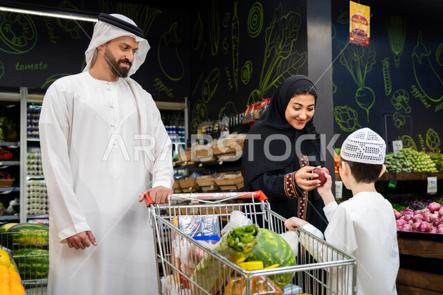 متعة التسوق والتبضع مع الأبناء في السوبرماركت، عائلة عربيه خليجيه اماراتية تتسوق في قسم الخضراوات بالسوبرماركت،ام عربيه خليجيه اماراتيه تقوم باعطاء ابنها ثمرة تفاح حمراء اللون، اختيار الفاكهه الطازجه المحليه الوطنيه الاماراتيه