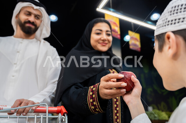 ايماءات تدل على السعادة، زوجان عربيان خليجيان اماراتيان يرتديان الزي التقليدي يقومان بقضاء أوقات ممتعة بالتسوق واختيار اللوازم المنزليه برفقه ابنهم، ام اماراتية تقوم باعطاء ولدها التفاح الاحمر، مساعدة وتعليم الأبناء الشراء من الماركت