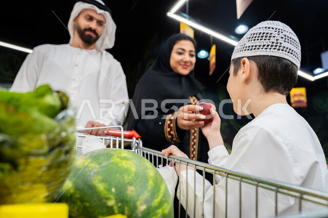 التعاون في شراء السلع والمواد الغذائيه المختلفة في السوبرماركت، زوجان عربيان خليجيان اماراتيان يرتديان الزي التقليدي يقومان بقضاء أوقات ممتعة بالتسوق واختيار اللوازم المنزليه برفقه ابنهم، ام اماراتية تقوم باعطاء ولدها التفاح الاحمر، محاصيل زراعية وطنية اماراتيه