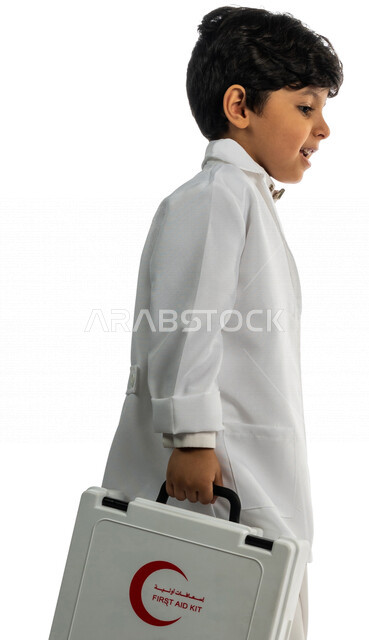 بورتريه بمنظر جانبي لفتى عربي خليجي سعودي ، يحمل بيده حقيبة الإسعافات الأولية ، يقوم بالسير بمفهوم لغة الجسد المختلفة ، يرتدي زي الطبيب ، الطبيب الصغير ، يحلم بأن يصبح طبيب ، المهنة المستقبلية ، مفهوم الطب والرعاية الصحية ، خلفية بيضاء
