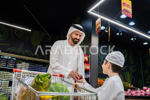 Family participation in purchasing household supplies and needs, an Emirati Gulf Arab man wearing a white kandura and ghutra accompanied by his son shopping in the fruits and vegetables section of the supermarket, enjoying shopping, local Emirati agricultural crops