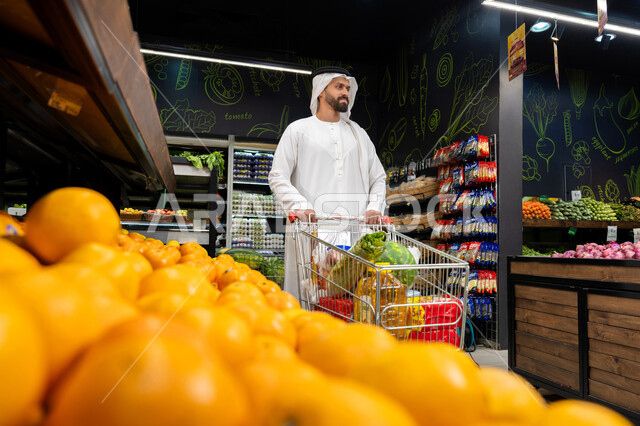 الاستمتاع بالتسوق و شراء الاحتياجات المنزلية والسلع والمواد الغذائية المختلفة، رجل عربي خليجي اماراتي يرتدي كندورة وغترة بيضاء يتبضع في قسم الفواكه والخضروات عند رف البرتقال، التسوق داخل السوبرماركت، سلة مشتريات ممتلئة بمستلزمات المطبخ، محاصيل زراعية اماراتية