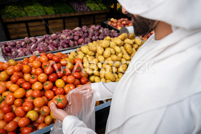 Having a good time shopping in the markets and choosing snacks and healthy foods, fresh colorful vegetables, buying household needs from the fruit and vegetable section in stores, a close-up of the hand of an Emirati Gulf Arab man wearing a kandura picking tomatoes, Emirati national agricultural crops