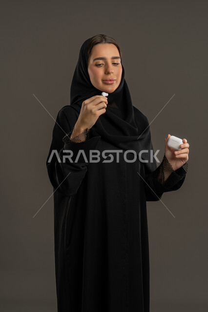 The concept of technology and communication, a modern Bluetooth headset connected to a mobile device, a portrait of an Arab Gulf Emirati woman wearing an abaya and hijab holding a wireless earphone in her hand, a Saudi woman using accessories for modern technical devices, black background.