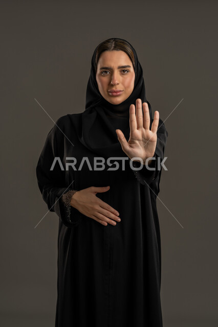 الأمر بالتوقف عن فعل شيء ما، بورتريه لامرأه عربيه خليجيه امارتية  ترتدي العباءة السوداء والحجاب ترفع يدها بإيماءات النهي، سيدة سعودية تنظر الى الكاميرا بتعابير الرفض والامتناع  ،الشعور بالإنزعاج والغضب، خلفية سوداء .