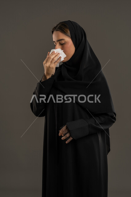 استعمال المناديل الورقية أثناء العطس، الإصابة بفيروس كورونا كوفيد19، بورتريه لامرأه عربيه خليجيه اماراتية  ترتدي العباءة السوداء والحجاب، إيماءات وجه ويدين تدل على العطاس، سيدة سعودية تشعر بالتعب والمرض ، خلفية سوداء .
