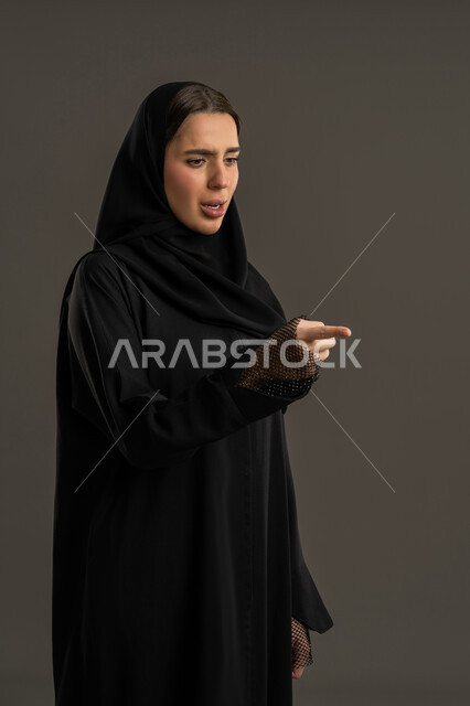 الاشارة الى شئ ما بتعابير الغضب ، بورتريه لامرأة عربية خليجية اماراتية ترتدي العباءة السوداء والحجاب ، سيدة سعودية تشير باصبع السبابة تجاه هدف ما ، بإيماءات وحركات يدين التوجيه لمكان معين ، خلفية سوداء.