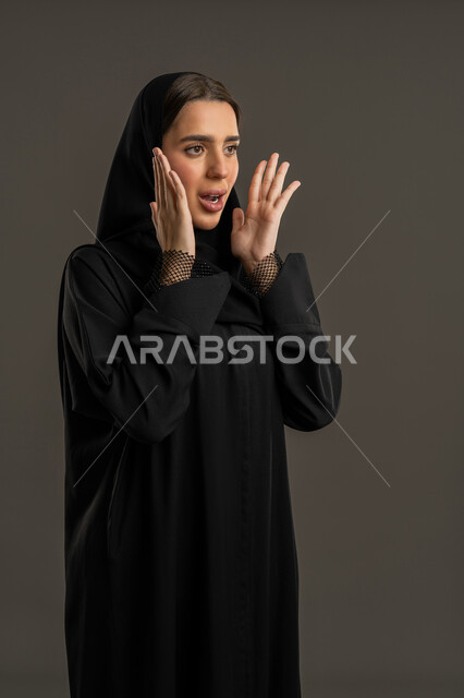 ايماءات الدهشة  لسماع خبر مفاجئ، التعجب من حصول أمر ما، بورترية مقرب لامرأة عربية خليجية اماراتية ترتدى العباءة السوداء والحجاب تنظر إلى شئ ما بتعابير الصدمة ،سيدة سعودية تضع يدها على وجهها بايماءات الذهول ، خلفية سوداء .