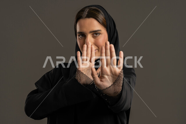 مد اليدين نحو الأمام في محاولة للدفاع عن النفس،  مفهوم الخوف والقلق ،  بورتريه مقرب لامراة عربية خليجية إماراتية ترتدي العباءة السوداء والحجاب تنظر إلى الكاميرا بإيماءات الرهبة، ترك مسافة أمان وطلب التوقف،سيدة سعودية ترفع يدها نحو وجهها,، خلفية سوداء .