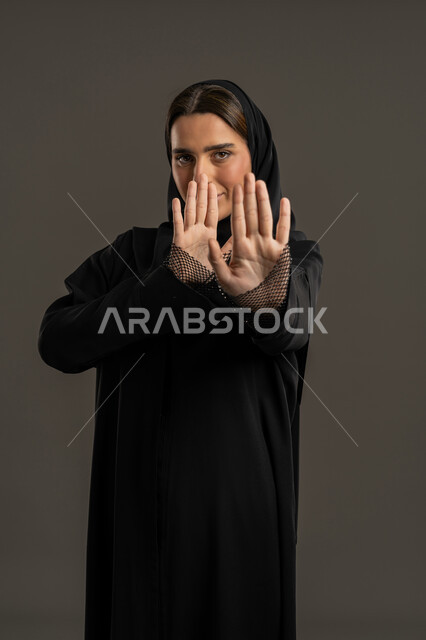 الذعر والخوف من شيء ما، مد اليدين نحو الأمام في محاولة للدفاع عن النفس، بورتريه لامراة عربية خليجية إماراتية ترتدي العباءة السوداء والحجاب تنظر إلى الكاميرا بإيماءات الرهبة، ترك مسافة أمان وطلب التوقف،سيدة سعودية ترفع يدها نحو وجهها,، خلفية سوداء .