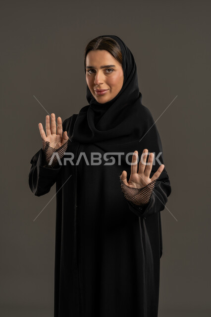 ترك مسافة أمان وطلب التوقف ، بورتريه لامراة عربية خليجية إماراتية ترتدي العباءة السوداء والحجاب تنظر إلى الكاميرا بإيماءات الرهبة، ،سيدة سعودية ترفع يدها في محاولة للدفاع عن النفس , الذعر والخوف من شيء ما ، خلفية سوداء .