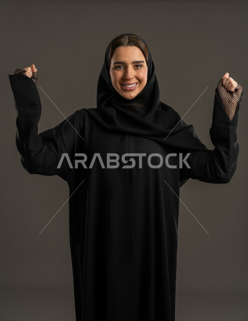 الفرح بالنصر والفوز، تحقيق الأهداف والنجاح، بورترية لامراه عربية إماراتية ترتدي العباءة السوداء والحجاب تنظر إلى الكاميرا بتعابير السرور والسعادة، بورتريه مقرب لامرأة خليجية سعودية محجبة مبتسمة ترفع قبضة يديها بإيماءات الحماس والانتصار ، خلفية سوداء .