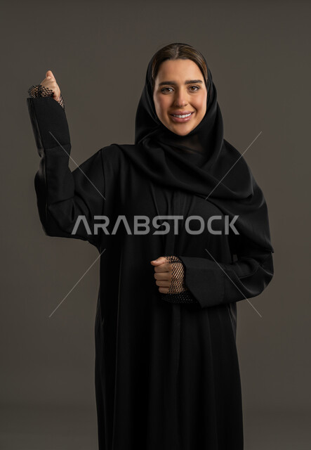 استعراض القوة البدنية، عرض عضلات اليدين، نشاط بدني للمحافظة على صحة الجسم، بناء العضلات وتقويتها، بورتريه لامرأة عربية خليجية اماراتية ترتدي العباءة السوداء والحجاب ،ترفع يدها بكفة مقبوضة،سيدة سعودية تنظر الى الكاميرا بايماءات السعادة ، خلفية سوداء.