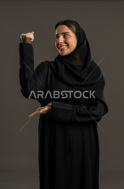 بناء العضلات وتقويتها، نشاط بدني للمحافظة على صحة الجسم، استعراض القوة البدنية ،عرض عضلات اليدين، بورتريه لامرأة عربية خليجية اماراتية ترتدي العباءة السوداء والحجاب ، سيدة سعودية ترفع يدها بكفة مقبوضة بايماءات السعادة ، خلفية سوداء.