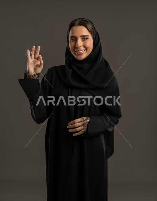 السرور والسعادة بإنجاز المهام، بورتريه لامرأة عربية خليجية اماراتية مبتسمة ترتدى العباءدة السوداء والحجاب تنظر إلى الكاميرا، سيدة سعودية ترفع يدها بعلامة الجودة والامتياز، الدقة في تحقيق أهداف العمل, خلفيه سوداء .