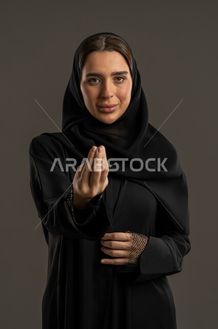 الاشارة بإيماءات الصبر والتأن ، إمهال بعض الوقت للقيام بشئ ما، التوعد والتهديد لشخص ما، مفهوم الصبر والتريث، بورتريه مقرب لامرأة عربية خليجية اماراتية ترتدي العباءة السوداء والحجاب تنظر الى الكاميرا، سيدة سعودية ترفع يدها بتعابير طلب الإنتظاروالتمهل، خلفية سوداء.