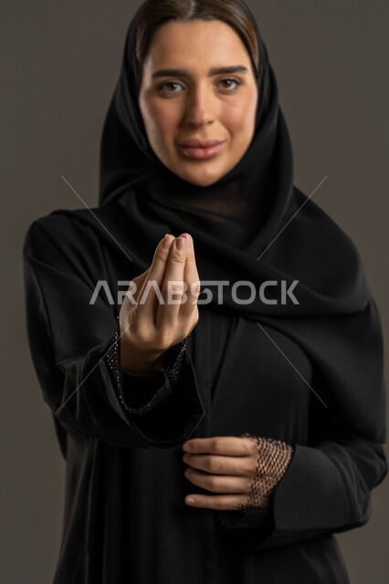 مفهوم الصبر والتريث، التوعد والتهديد لشخص ما، بورتريه مقرب لامرأة عربية خليجية اماراتية ترتدي العباءة السوداء والحجاب تنظر الى الكاميرا، سيدة سعودية ترفع يدها بتعابير طلب الإنتظار والتمهل، خلفية سوداء.