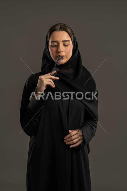 Weak and nearsightedness, the concept of health care and appearance, interest in fashion and elegance, a portrait of an Emirati Gulf Arab woman wearing an abaya and hijab, holding eyeglasses in her hand with an expression of self-confidence, a Saudi woman looking at something, black background.