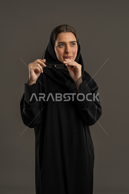 مفهوم العناية بالصحة والمظهر الخارجي، ضعف وقصر النظر، الاهتمام بالموضة والأناقة, بورتريه لامرأة عربية خليجية إماراتية ترتدي العباءة والحجاب تمسك بيدها النظارات الطبية بتعابير الثقة بالنفس, سيدة سعودية تنظر الى شئ ما بإيماءات السعادة, خلفية سوداء .