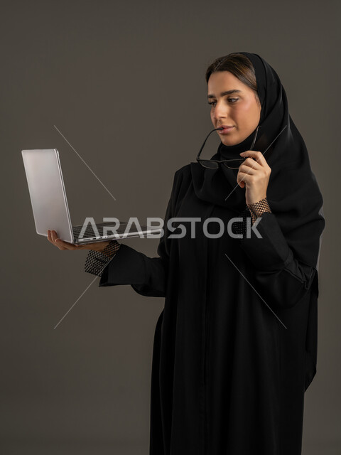مهن ووظائف نسائية ، ايماءات الاندماج والتركيز لإنجاز المهام ، ادارة العمل عن بعد ، إستخدام جهاز الحاسوب المحمول ، بورتريه لامراة عربية خليجية اماراتية تمسك نظاره بيدها و جهاز اللابتوب ، إستخدام أجهزة تقنية حديثة ، سيدة سعودية تقوم بتصفح الخدمات والتطبيقات عبر الانترنت ، مفهوم الإعلانات والتسويق الرقمي الإلكتروني ، خلفية سوداء .