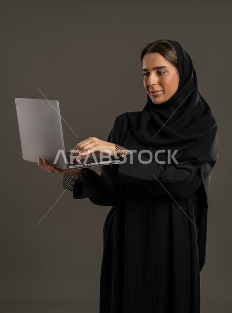 Gestures of integration and focus to accomplish tasks, remote work management, using a laptop, portrait of an Arab Gulf Emirati woman using a laptop, using modern technology devices, a Saudi woman browsing online services and applications, the concept of advertising and digital electronic marketing, black background.