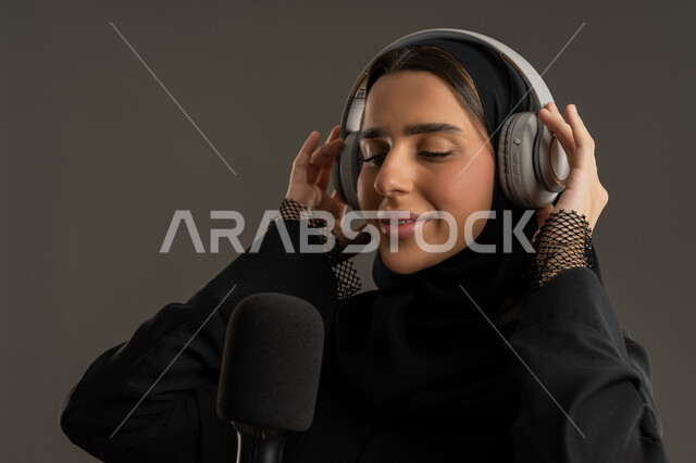 دمج التقنيات الحديثة في الحياة العملية ، الاذاعة الصوتية والغناء والطرب ، اجراء بث صوتي مباشر (بودكاست) عبر برامج التواصل الاجتماعي ، بورتريه مقرب لامرأة عربية خليجية اماراتية مبتسمة ترتدي الحجاب والعباءة،، معدات وأجهزة تسجيلات الصوت، سيدة سعودية ترتدي سماعات الرأس وتستمتع بتقديم البرامج ، خلفية سوداء .