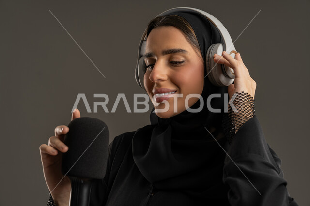 الاذاعة الصوتية والغناء والطرب ، اجراء بث صوتي مباشر (بودكاست) عبر برامج التواصل الاجتماعي ، بورتريه مقرب لامرأة عربية خليجية اماراتية مبتسمة ترتدي الحجاب والعباءة،، معدات وأجهزة تسجيلات الصوت، سيدة سعودية ترتدي سماعات الرأس وتستمتع بتقديم البرامج ، خلفية سوداء .