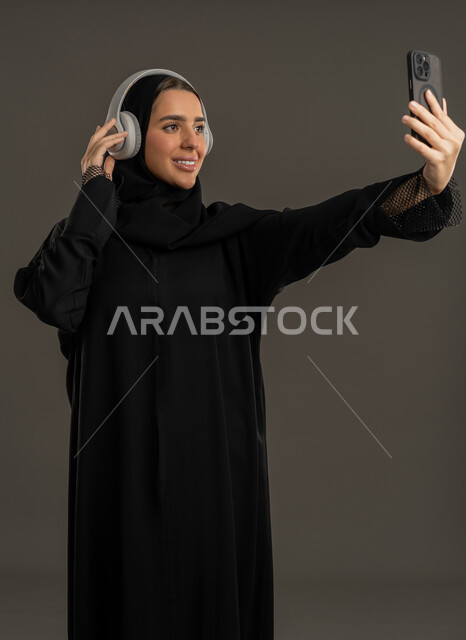 برامج وتطبيقات الجوال المتنوعة، توثيق اللحظات السعيدة، اجراء مكالمات الفيديو ، التواصل مع الأهل والأصدقاء ، دمج التكنولوجيا المتطورة في الحياة اليومية، بورتريه لامرأة عربية خليجية اماراتية ترتدي العباءة السوداء والحجاب تحمل بيدها الهاتف المحمول وتنظر إليه بإيماءات السرور ، سيدة سعودية تلتقط صورة تذكارية وتمسك سماعات الرأس ، خلفية سوداء .