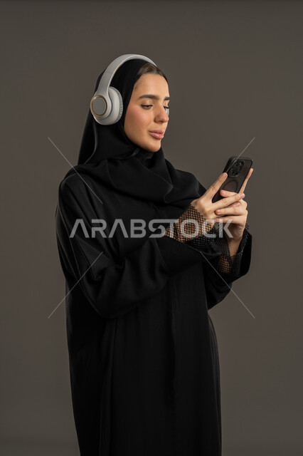 مفهوم الإصغاء والإستماع ،ايماءات وجه ويدين تدل على السعادة والاندماج، الاستمتاع بالموسيقى في أوقات الفراغ ,  بورتريه من الجانب مقرب لإمرأة عربية اماراتية خليجية ترتدي العباءة السوداء والحجاب تضع  سماعة الراس  ، سيدة سعودية تمسك الجوال بيدها ،استخدام التقنيات المتطورة ، خلفية سوداء .