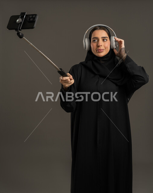استخدام الاجهزة الحديثة ، التقاط الصور التذكارية الجميلة، اجراء مكالمات الفيديو ، التواصل مع الأهل والأصدقاء ، دمج التكنولوجيا المتطورة في الحياة اليومية، بورتريه لامرأة عربية خليجية اماراتية ترتدي العباءة السوداء والحجاب تحمل بيدها الهاتف المحمول وتنظر إليه بإيماءات السرور ، سيدة سعودية تلتقط صورة تذكارية  بواسطة عصا السيلفي ، خلفية سوداء .