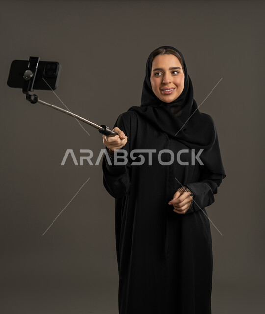 التقاط الصور التذكارية الجميلة، اجراء مكالمات الفيديو ، التواصل مع الأهل والأصدقاء ، دمج التكنولوجيا المتطورة في الحياة اليومية، بورتريه لامرأة عربية خليجية اماراتية ترتدي العباءة السوداء والحجاب تحمل بيدها الهاتف المحمول وتنظر إليه بإيماءات السرور ، سيدة سعودية تلتقط صورة تذكارية  بواسطة عصا السيلفي ، خلفية سوداء .