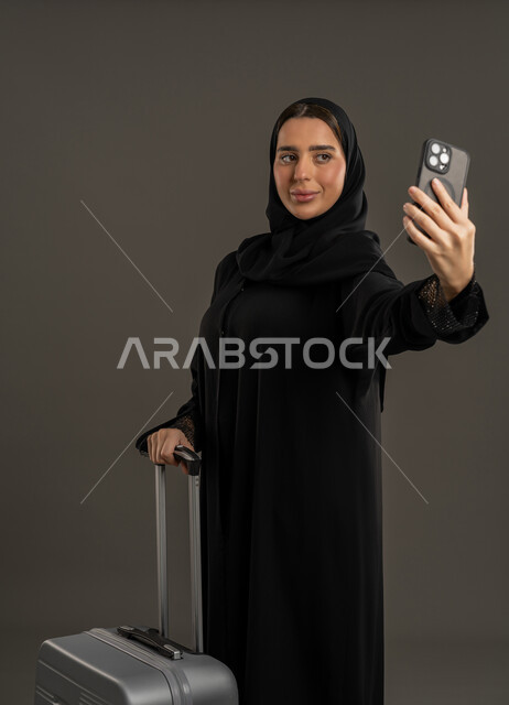 دمج التكنولوجيا المتطورة في الحياة اليومية، إجراء مكالمات الفيديو والتواصل مع الأهل والأصدقاء، بورتريه لامرأة عربية خليجية اماراتية ترتدي العباءة السوداء والحجاب تحمل بيدها الهاتف المحمول وتنظر إليه بإيماءات السرور ، سيدة سعودية تمسك بيدها شنطة السفر، التقاط صور السيلفي، خلفية سوداء .