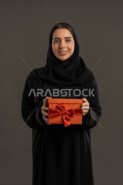 تقديم الهدايا فى الأعياد والمناسبات والمفاجآت السعيدة، بورتريه لامرأة عربية خليجية اماراتية ترتدي العباءة السوداء والحجاب تحمل بيديها علبة مغلفة، سيدة سعودية تنظر الى الكاميرا بإيماءات السرور ، خلفية سوداء .