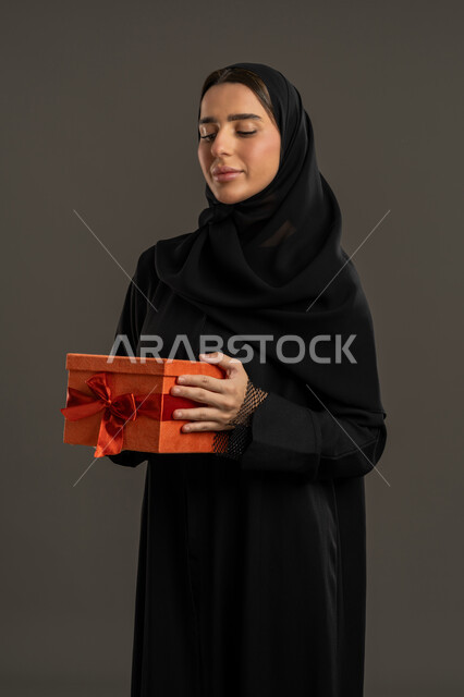 Expressing love and appreciation, gestures of joy when receiving gifts, happiness in giving gifts and congratulations, happy holidays and occasions, portrait of an Arab Gulf Emirati woman wearing a black abaya and hijab holding a wrapped box in her hands, a Saudi woman looking at the gift with an expression of pleasure, black background.