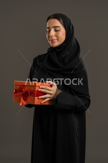 مفهوم المفاجآت السعيدة، ايماءات الفرح عند الحصول على الهدايا، السعادة بتقديم الهدايا والتهنئات، أعياد ومناسبات سعيدة, بورتريه لامرأة عربية خليجية اماراتية ترتدي العباءة السوداء والحجاب تحمل بيديها علبة مغلفة، سيدة سعودية تنظر الى الهدية بتعابير السرور، خلفية سوداء .
