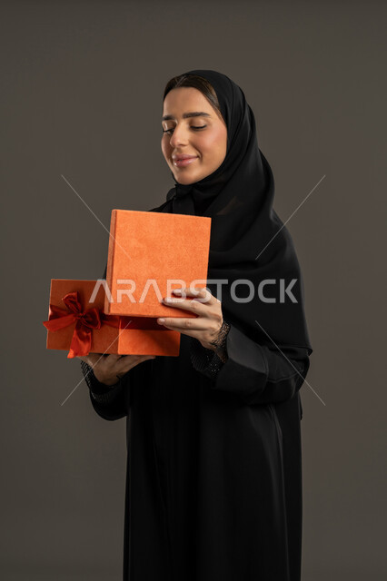 الحصول على الهدايا والتهنئة ، أعياد ومناسبات ومفاجآت سعيدة، ايماءات الفرح والسعادة, بورتريه لامرأة عربية خليجية اماراتية ترتدي العباءة السوداء والحجاب ، تفتح علبة برتقالية مغلفة ، سيدة سعودية تنظر الى الهدية بتعابير السرور، خلفية سوداء .