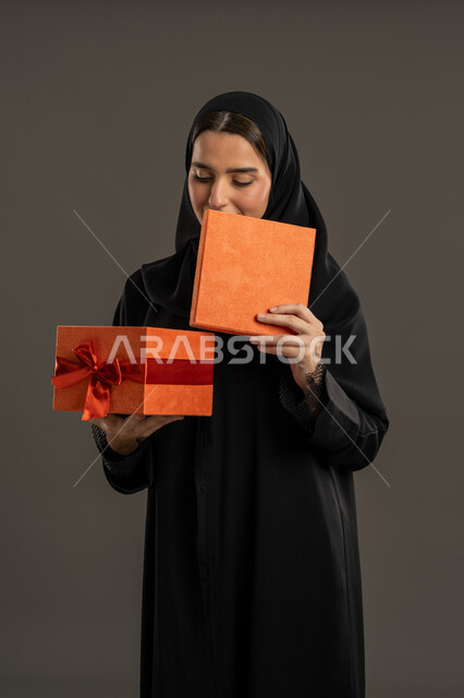 Holidays, occasions and happy surprises, gestures of joy and happiness by offering gifts and congratulations, portrait of an Arab Gulf Emirati woman wearing a black abaya and hijab, opening a wrapped orange box, a Saudi woman looking at the gift with an expression of pleasure, black background.