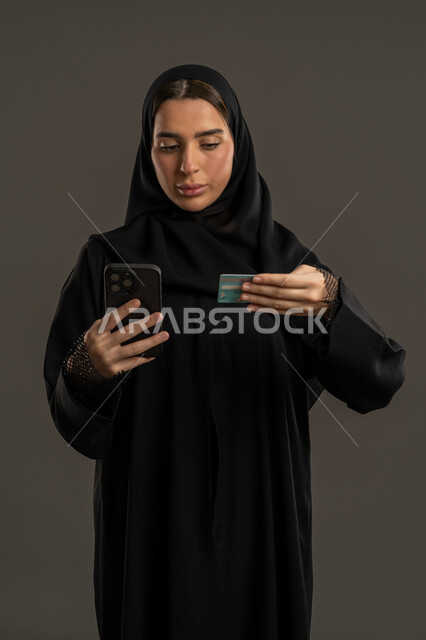 إمكانية تسديد الفواتير عن طريق بطاقة الإئتمان البنكية، استخدام تطبيقات الجوال للدفع والشراء، بورتريه لإمرأة عربية خليجية اماراتية مبتسمة ترتدي العباءة السوداء والحجاب تمسك بيدها بطاقة الصراف، سيدة سعودية تنظر الى البطاقة الالكترونية  بإيماءات التركيز، خلفية سوداء.