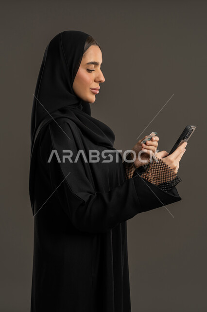 استخدام تطبيقات الجوال للدفع والشراء، إمكانية تسديد الفواتير عن طريق بطاقة الإئتمان البنكية، بورتريه من الجانب لإمرأة عربية خليجية اماراتية مبتسمة ترتدي العباءة السوداء والحجاب تمسك بيدها بطاقة الصراف، سيدة سعودية تنظر الى الهاتف وتستخدم التقنيات الحديثة المتطورة ، خلفية سوداء.