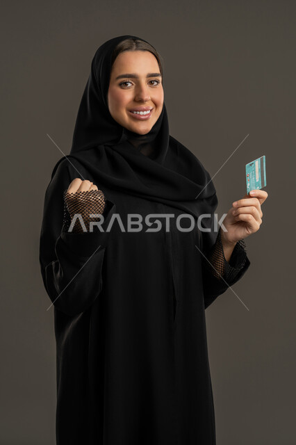 إمكانية تسديد الفواتير بالبطاقة الإلكترونية، الشراء والدفع عن طريق بطاقة الإئتمان البنكية، بورتريه لإمرأة عربية خليجية اماراتية مبتسمة ترتدي العباءة السوداء والحجاب تمسك بيدها بطاقة الصراف، سيدة سعودية تنظر الكاميرا بإيماءات وتعابير السعادة والفرح ، خلفية سوداء.