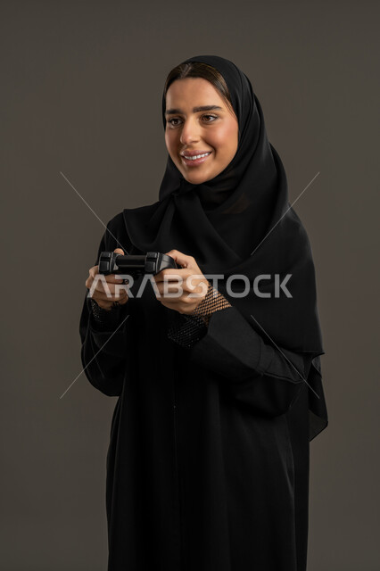 Digital entertainment world, video game entertainment, electronics and modern devices, portrait of an Arab Gulf Emirati woman wearing a black abaya and hijab, holding a joystick in her hand with focus and immersion, a Saudi woman looking at the camera with happy gestures, enjoying a fun and exciting entertainment experience, black background.