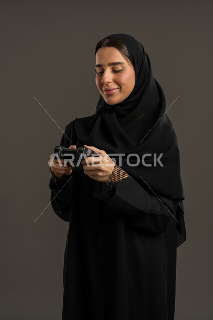 الحماس بالعالم الرقمي الالكتروني الحديث والمتطور، الألعاب والأجهزة الالكترونية الحديثة ، اللعب و التسلية بالعاب الفيديو ، بورترية لامراة عربية خليجية إماراتية ترتدي العباءة السوداء والحجاب ، تمسك بيدها عصا التحكم بتركيز وتنظر اليه , سيدة سعودية تستمتع بتجربة ترفيهية مثيرة ،خلفية سوداء .