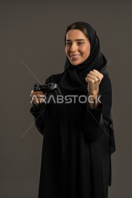 مفهوم الاندماج والتفاعل ، الألعاب الالكترونية والأجهزة الحديثة ، العالم الرقمي الترفيهي ، اللعب و التسلية بالعاب الفيديو ، بورترية لامراة عربية خليجية إماراتية ترتدي العباءة السوداء والحجاب ، تمسك بيدها عصا التحكم بتركيز , سيدة سعودية ترفع قبضة يدها بايماءات الفوز ، الاستمتاع بتجربة ترفيهية مثيرة ،خلفية سوداء .