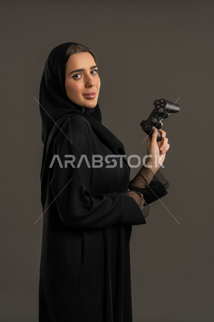 شغف العاب الفيديو الإلكترونية ، مفهوم تمكين المرأة في مجالات الترفيه والتقنية ، بورترية من الجانب لامراة عربية خليجية إماراتية ترتدي العباءة السوداء والحجاب ، تمسك بيدها عصا التحكم بتركيز واندماج, العالم الرقمي ، سيدة سعودية تنظر الى الكاميرا بايماءات السعادة ، الاستمتاع بتجربة ممتعة ومثيرة ، خلفية سوداء .