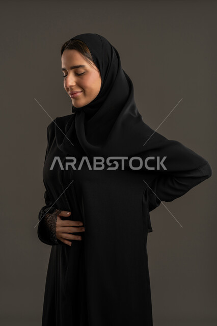 الحاجة لوضع خطة علاجية ، بورتريه لامرأة عربية خليجية امارتية ترتدي العباءة السوداء والحجاب ، تضع يديها على ظهرها و بطنها, ، أعراض الحمل ، سيدة سعودية تحتاج للمساعدة , خلفية سوداء .