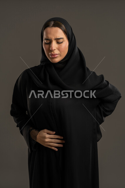 إيماءات يدين بالألم و الوجع  ، بورتريه لامرأة عربية خليجية امارتية ترتدي العباءة السوداء والحجاب ، تضع يديها على ظهرها و بطنها, ، أعراض الحمل ، سيدة سعودية تحتاج للمساعدة وطلب العلاج, خلفية سوداء .